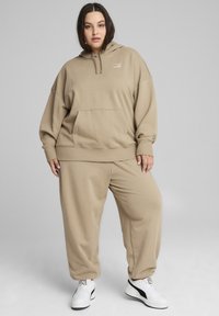 Beige huppari, jossa on huppu ja suuri etutasku, yhdistettynä löysiin sweatpants-housuihin. Päällä valkoiset lenkkarit, joissa on mustia yksityiskohtia.