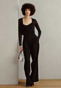 Black jumpsuit with long, sheer sleeves, a wrap design, and wide-leg fit; paired with a silver handbag and high heels. Smooth texture.