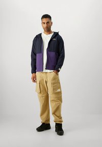 Meeste jope tumesinises ja lillakas toonis, millel on esil tõmblukk, küljetaskud ja kapuuts. Sobitatud beežide cargo-pükstega ning mustade kingadega.