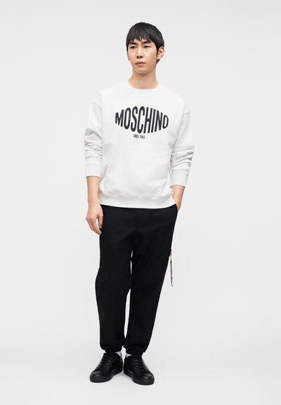 Λευκό φούτερ με μαύρο λογότυπο "MOSCHINO", συνδυασμένο με μαύρο παντελόνι και μαύρα αθλητικά παπούτσια. Απλό φόντο, το μοντέλο στέκεται με τα χέρια στις τσέπες.