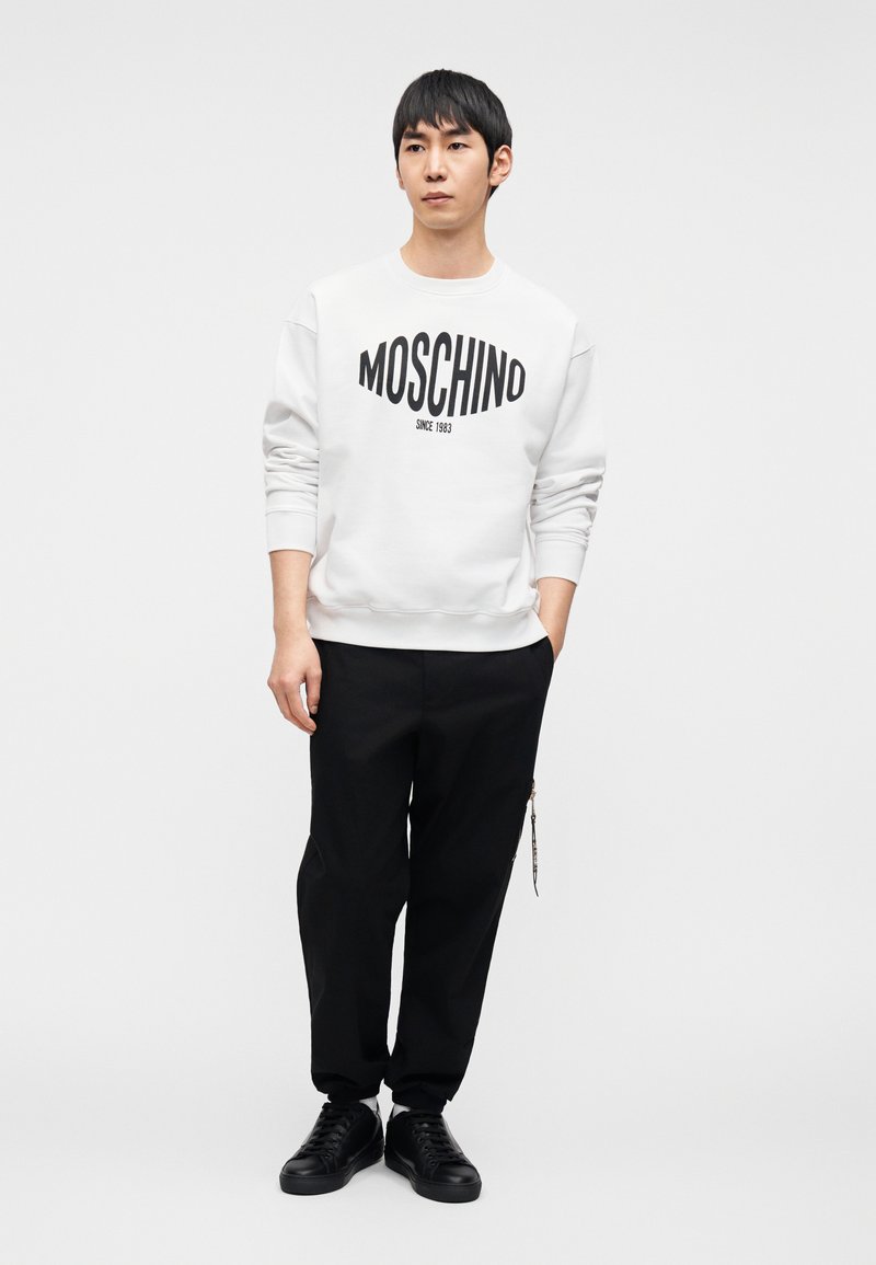 Sweat-shirt blanc avec logo "MOSCHINO" noir, associé à un pantalon noir et des baskets noires. Fond uni, le modèle pose les mains dans les poches.