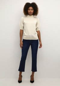 Haut en tricot beige clair avec un col montant, des manches ballon, associé à un jean noir cropped et des chaussures à talons hauts noires. Texture lisse.