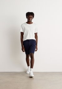 Hvit bomull T-skjorte med trykket tekst, parret med marineblå shorts. Modellen har på seg hvite sneakers og står på en beige betonggulv.