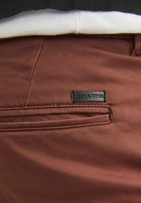 Burgundowe spodnie o gładkiej fakturze, z tylną kieszenią i czarną metką z napisem "JACK & JONES" przyszytą z tyłu.