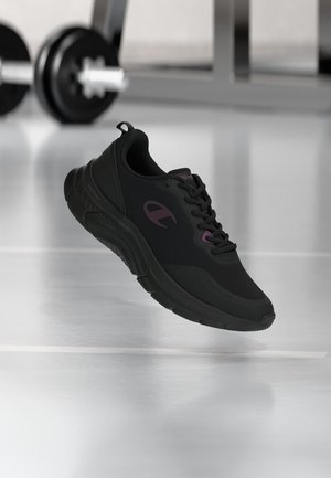 Sort atletiksneaker med lilla logo og accenter, svævende i luften i et fitnesscenter med slørede håndvægte i baggrunden.