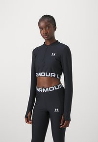 Crni sportski crop top dugih rukava s polu-zatvaračem, uparen s crnim tajicama visokog struka koje imaju kontrastni logo na pojasu.