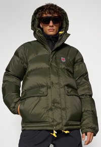 Veste en duvet verte avec une finition texturée, capuche, boutons à l'avant et deux poches ; présente un patch logo et des accents jaunes sur les cordons.