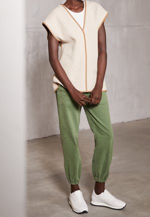 Person trägt ärmelloses, beige Stricktop mit braunem Besatz, grüne Hose mit elastischem Saum und weiße Sneaker, steht auf Betonboden.