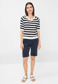 Stripet V-hals sweater i marineblå og hvid, parret med marineblå shorts. Shortsene har rullede kanter, og sandalerne er hvide med en spænde.