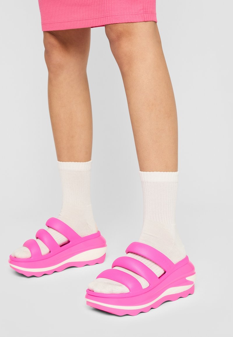 Sandales compensées roses avec trois larges brides et une semelle blanche ondulée. Portées avec des chaussettes blanches côtelées. Texture lisse dans l'ensemble.