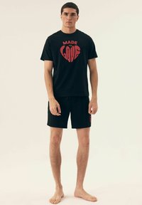 Czarny bawełniany T-shirt z wyraźnym czerwonym nadrukiem, na którym widnieje napis "STWORZONE DO KOCHANIA", zestawiony z pasującymi czarnymi szortami i bosymi stopami.