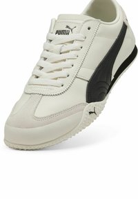Zapatilla de cuero blanca con acentos negros, lados texturizados y una puntera de gamuza. Presenta cordones redondos y una suela de goma con pequeñas protuberancias.
