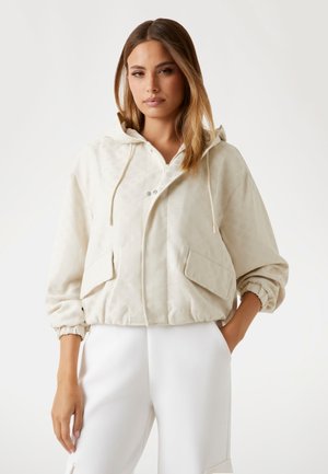 Cream hooded jacket with zip closure, front pockets, elastic cuffs, and a textured surface pattern. Paired with white trousers.