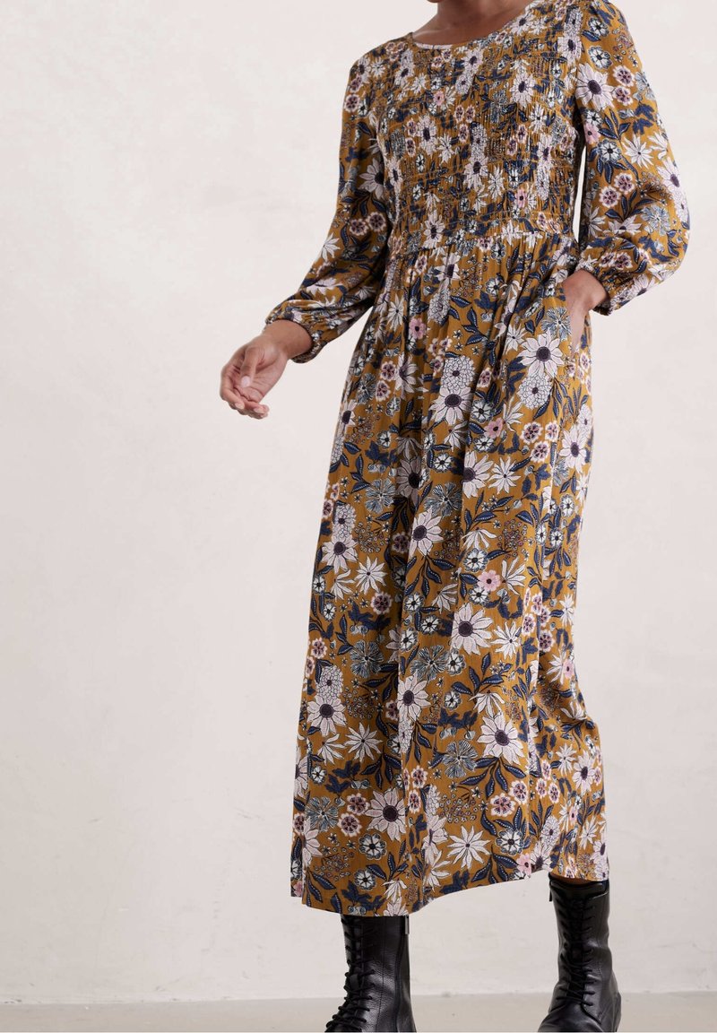 Femme portant une longue robe fleurie à fond moutarde et des bottes noires à lacets, une main dans la poche, debout contre un mur pâle.