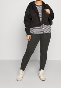 Svart kort hoodie med framfickor, randig skjorta, mörkgrå leggings och beige sneakers med ljusa snören. Slät textur genom hela plagget.
