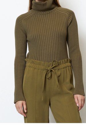 Femme portant un pull à col roulé côtelé olive et un pantalon taille haute assorti avec une taille à cordon, debout devant un fond blanc.