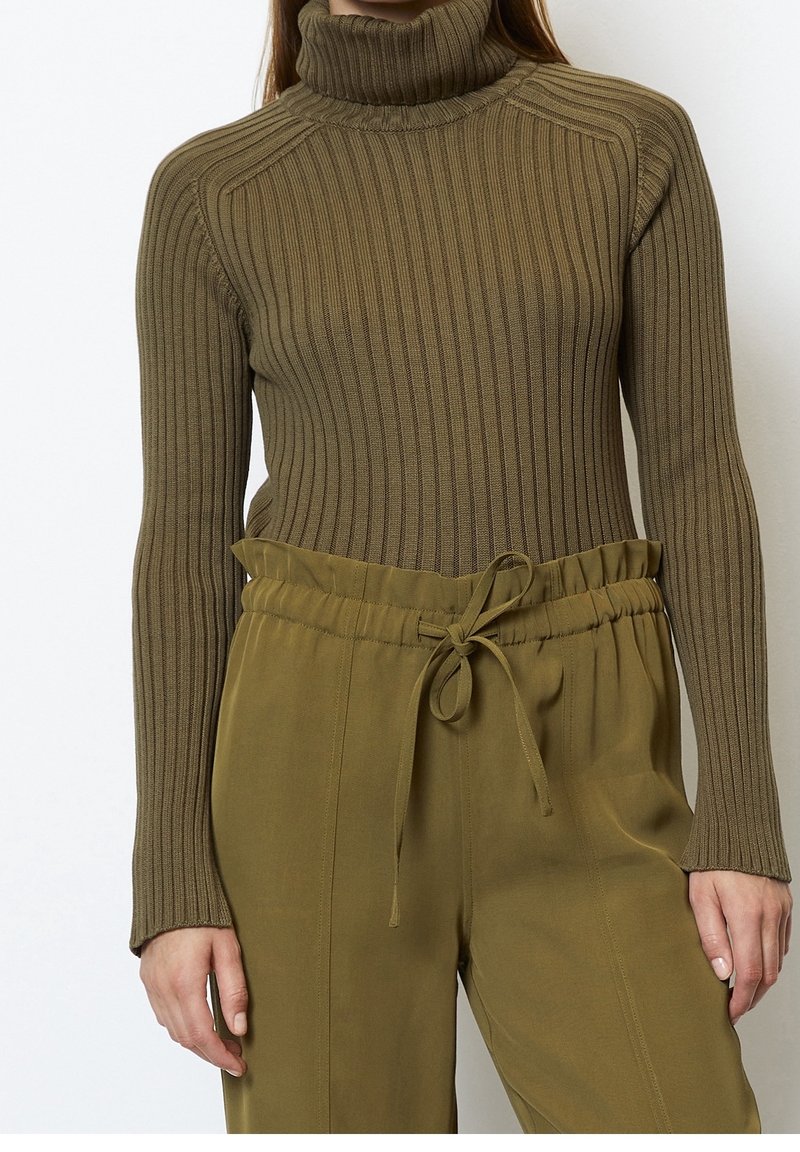 Femme portant un pull à col roulé côtelé olive et un pantalon taille haute assorti avec une taille à cordon, debout devant un fond blanc.
