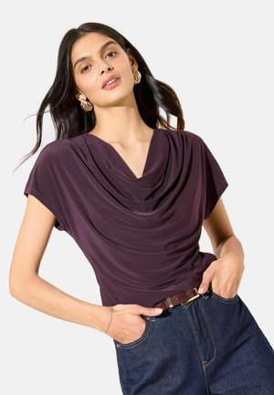Plum draped top with a V-neckline and short sleeves, made of smooth fabric. Paired with dark denim jeans and a brown belt.