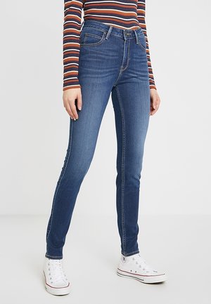 Jeans ajustados de denim azul oscuro; cuentan con un diseño clásico de cinco bolsillos, cintura a media altura y costuras claras. Combinados con zapatillas blancas.