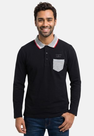 Homme souriant portant un polo noir à manches longues avec col et poche gris, debout, la main dans la poche d’un jean, sur fond blanc.