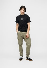 Νεαρός άνδρας που φοράει μαύρο γραφικό t-shirt, μπεζ παντελόνι cargo και μαύρα αθλητικά παπούτσια με λευκά κορδόνια, στέκεται με το ένα χέρι στην τσέπη.