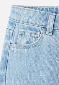 Material de denim albastru deschis cu o suprafață texturată, având un buzunar frontal, talie și accente decorative cu nituri.