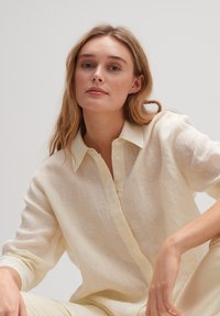 Light yellow, linen-blend shirt with a collared neckline, front buttons, and rolled sleeves; soft texture and subtle weave pattern.