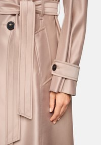 Hellbeiger Trenchcoat mit satinartigem Finish. Verfügt über einen Gürtel, einen knopfförmigen Ärmelabschluss und eine diagonale Tasche auf der linken Seite. Glatte Textur.