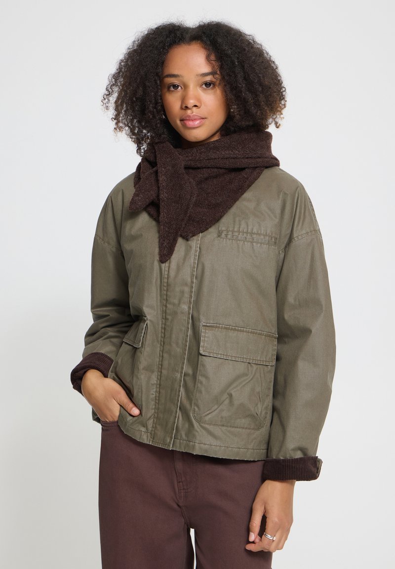 Jeune femme aux cheveux bouclés porte une veste verte, une écharpe marron et un pantalon marron, debout avec une main dans la poche sur un fond uni.