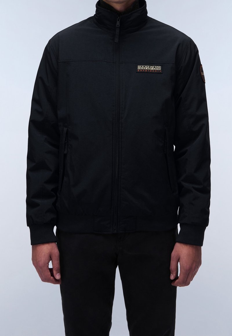 Homme portant une veste noire zippée avec le logo Napapijri Geographic sur la poitrine et un patch sur le bras gauche, debout sur un fond gris.