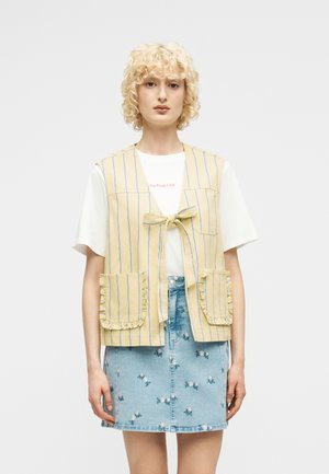 Personne blonde portant un gilet rayé jaune avec des poches avant par-dessus un t-shirt blanc et une jupe en jean bleu avec une petite broderie florale.