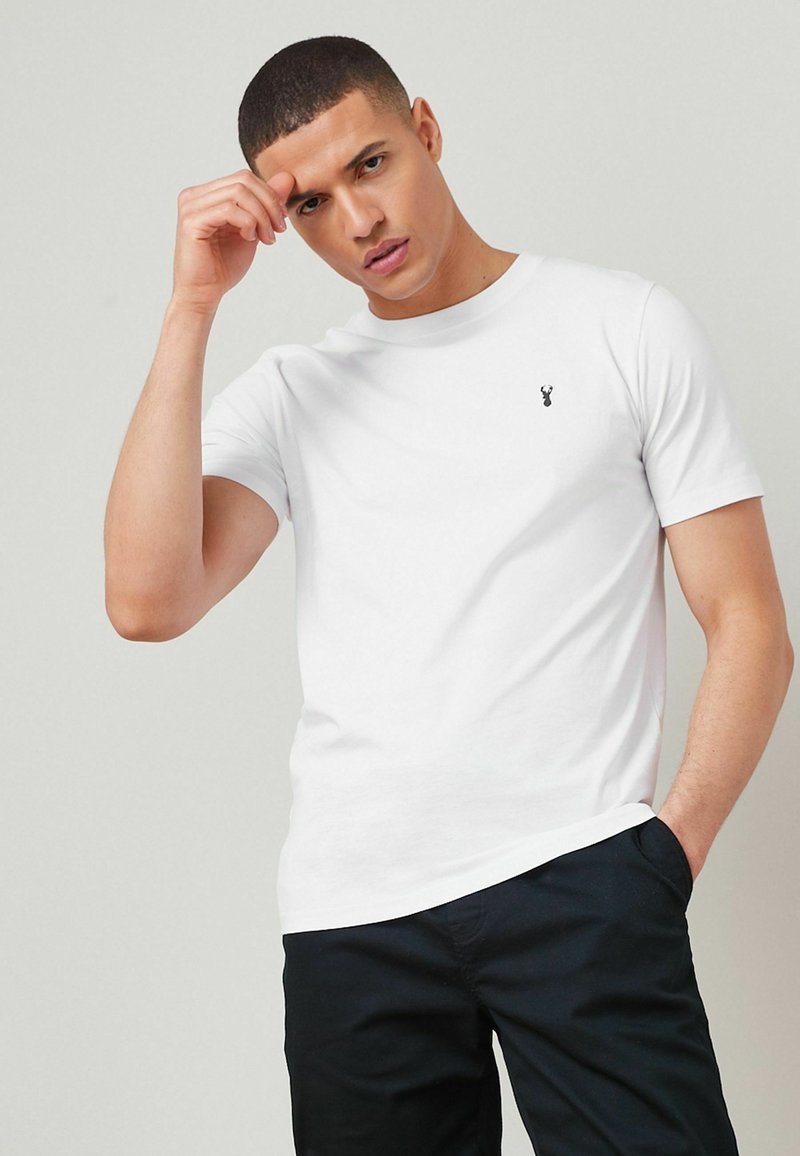Camiseta blanca de algodón con cuello redondo y mangas cortas, con un pequeño logo negro en el lado izquierdo del pecho, combinada con pantalones oscuros.