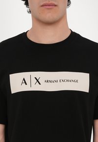Svart T-skjorte i myk bomull med et stort kremfarget rektangel hvor "A|X ARMANI EXCHANGE" er trykket med fet svart tekst. Rund hals-design.
