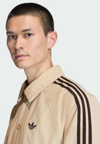 Beige lichtgewicht jack met een puntige kraag, een zwart driekleurig ontwerp langs de schouders en een klein zwart logo op de borst.