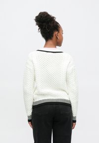 Hvid strikket trøje med tekstureret mønster, sort- og hvide stribede ribkanter ved kant og manchetter, båret med sorte jeans, set bagfra.