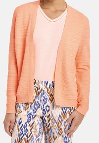 Vrouw in een gemêleerde perzikkleurige cardigan over een lichtroze V-hals top en gestreepte broek met abstracte ontwerpen in blauw, bruin en beige.