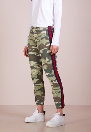 Personne portant un pantalon de jogging imprimé camouflage avec des bandes latérales rouges et bleues, un haut blanc à manches longues et des baskets blanches, debout sur un sol beige.