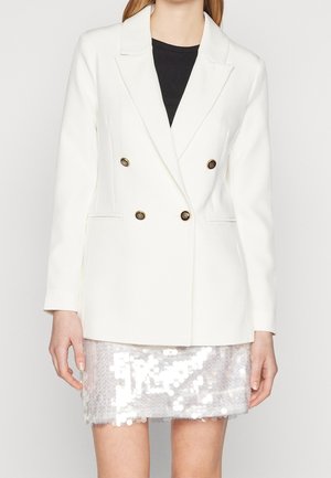 Kobieta ubrana w biały dwurzędowy blezer z złotymi guzikami na czarnym topie oraz krótką białą spódniczkę z cekinami.