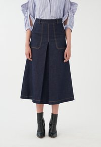 Falda de mezclilla con cintura alta, abertura frontal y costuras amarillas en contraste. Combinada con botas negras de tobillo con punta afilada.