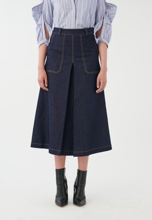 Jeansrock mit hoher Taille, vorderer Schlitze und kontrastierenden gelben Nähten. Kombiniert mit schwarzen Stiefeletten mit spitzer Schuhspitze.