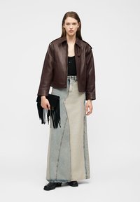 Γυναίκα που φοράει σκούρο καφέ δερμάτινο μπουφάν, μαύρο τοπ, μακρύ τζιν φούστα σε δύο τόνους μπλε και μπεζ, μαύρες μπότες, κρατώντας μαύρο τσαντάκι με φούντες.