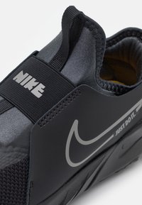 Baskets Nike noires avec une tige lisse, un design extensible, une sangle élastique et un logo bien en vue. La texture inclut des accents en mesh sur les côtés.