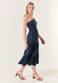 Marineblaues, ein-schultriges Kleid mit enganliegendem Oberteil und gestuftem, rüschendem Rock. Das Model trägt sandfarbene, Absatzsandalen mit dünnen Riemen.