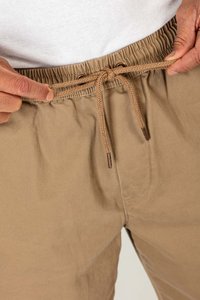 Pantalones de algodón beige con cinturilla elástica, que cuentan con un cordón de ajuste y puntas de metal. Textura suave con detalles de costura visibles en los laterales.
