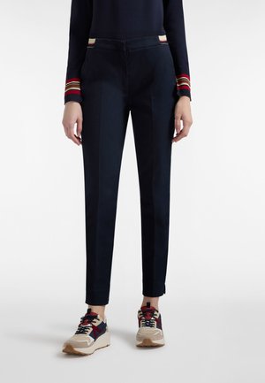 Pantaloni blu navy con una vestibilità su misura, caratterizzati da righe sui polsini e una texture liscia. Abbinati a sneaker beige e rosse.