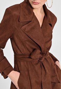 Brauner Wildleder-Trenchcoat mit tailliertem Schnitt, großen Revers und Seitentaschen. Glatte Textur und maßgeschneiderte Passform, die ein elegantes Design unterstreicht.