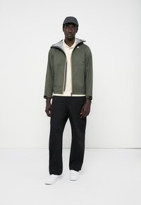 Grønn lett jakke med grå hette, glidelås og oransje detaljer; paret med en beige polo, svarte cargo-bukser og hvite joggesko.