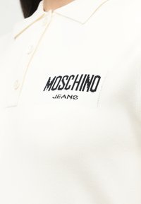 Λευκό πόλο πουκάμισο με τρία κουμπιά και κεντημένο μαύρο λογότυπο "MOSCHINO JEANS" στο αριστερό στήθος.