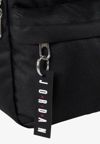 Črna tkaninska torba z teksturirano površino, ki ima srebrno zadrgo in črno etiketo z napisom "JORDAN" v krepko rdečih in belih črkah.