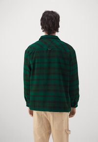 Camicia a scacchi verde e nera con colletto, maniche lunghe e orlo dritto. Dettagli notevoli includono un'etichetta ad anello vicino al colletto e una consistenza morbida.